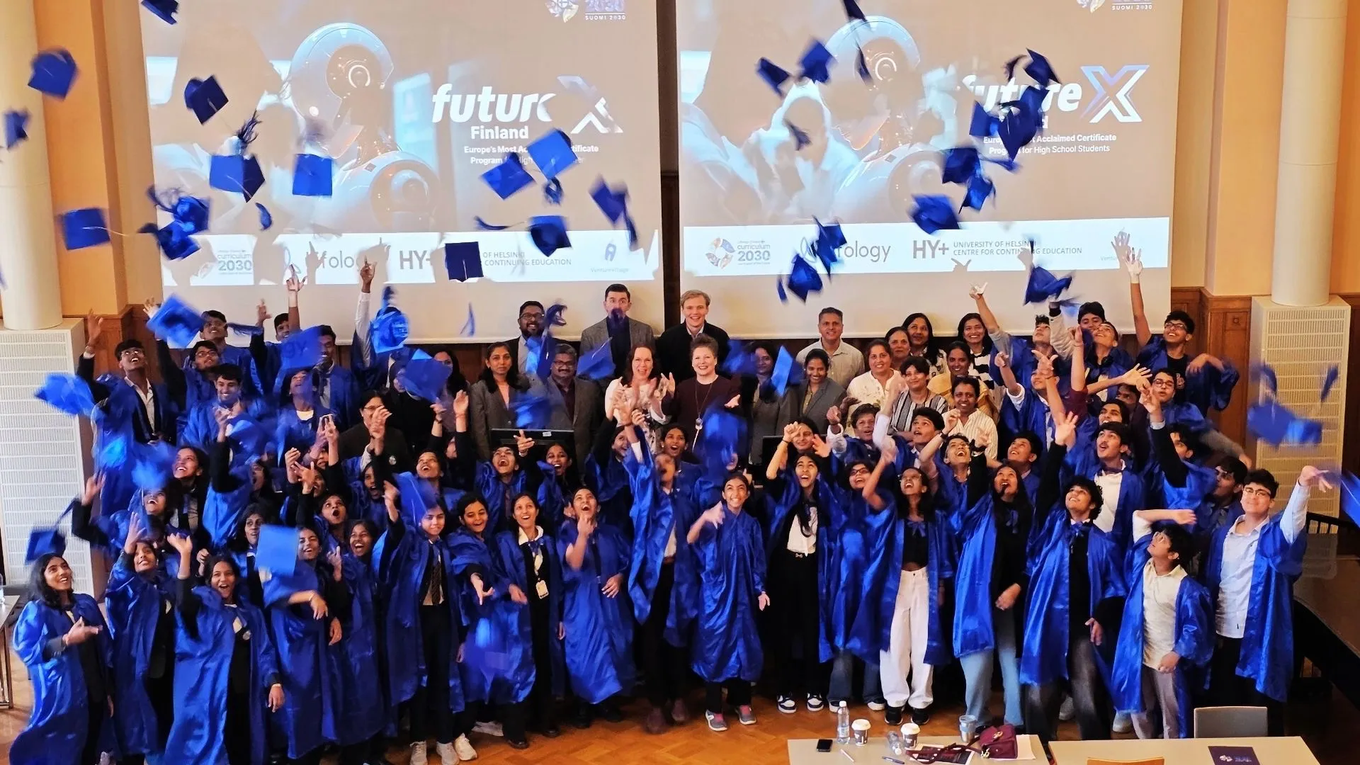 A large group of students in blue graduation gowns throwing their caps into the air in a hall.